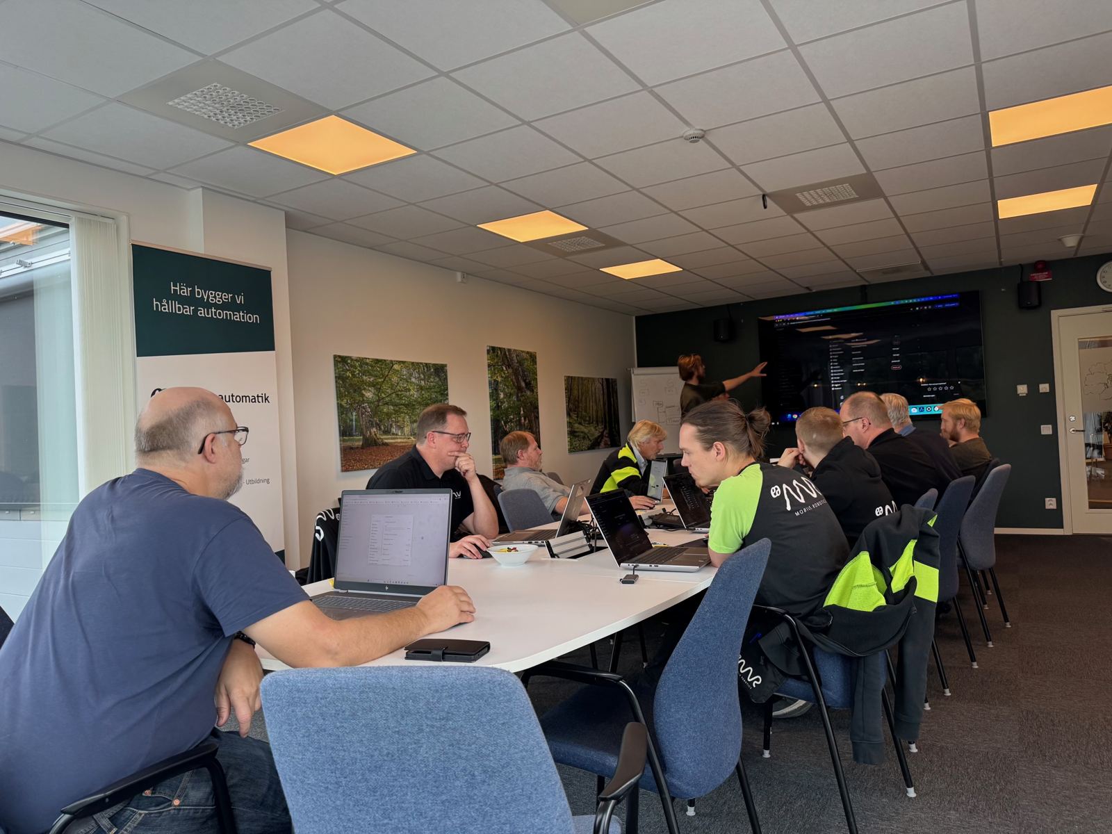Kursledare pekar på storskärmen medan deltagarna arbetar i laptops under en teknisk workshop